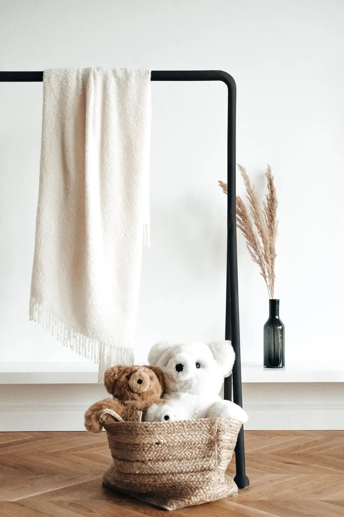 Two teddy bears in a woven basket with a blanket draped over a black rack on a wooden floor.