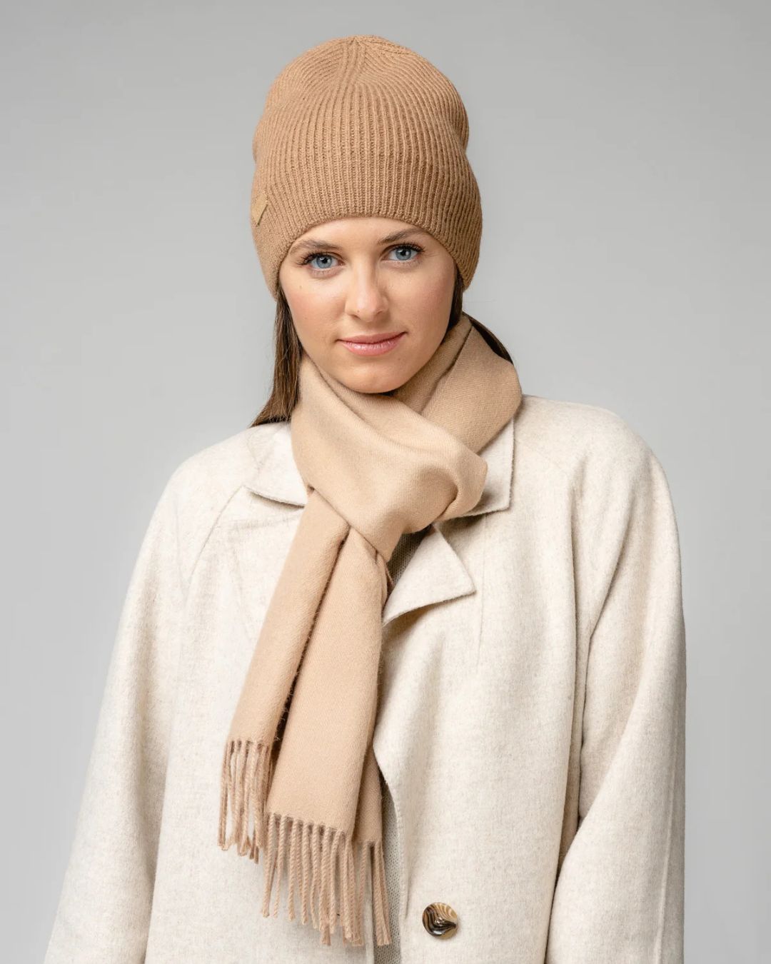 A woman wearing a ribbed knit beanie and a matching scarf in a warm beige/tan color, paired with a cream-colored coat, against a solid gray studio background.