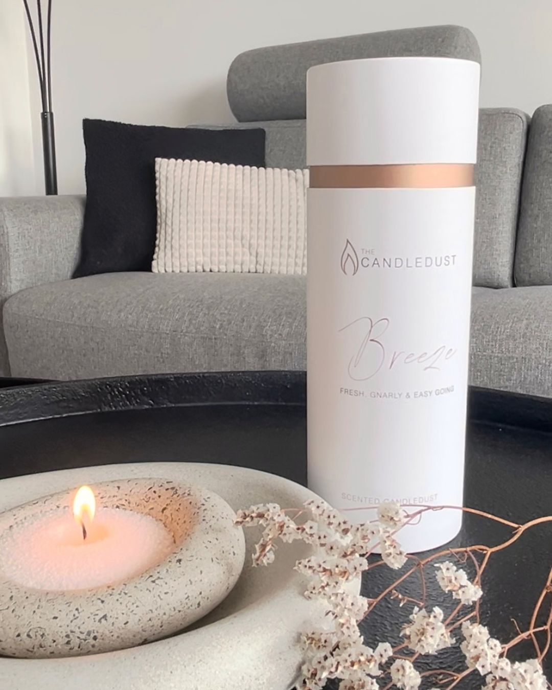 A white cylindrical CandleDust 'Breeze' fresh turmeric and baby body candle in a white cardboard sleeve, styled on a black tray next to a lit tealight candle in a speckled stone holder, with a gray sofa in the background.