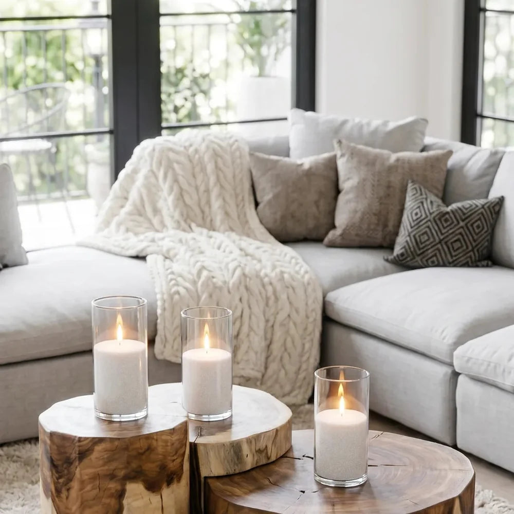 White pillar candles in clear glass holders on wooden side tables in a cosy Scandinavian living room with a cream knitted throw and soft neutral cushions